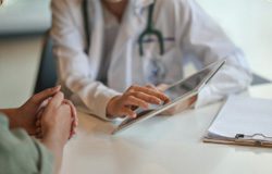 featured-appointments A doctor in a white coat uses a tablet during a consultation with a patient. The patient’s hands are clasped on the table, and a clipboard with papers is nearby.