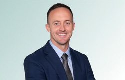 featured-mchugh Smiling man wearing a navy suit, light blue shirt, and gray tie, standing with arms crossed in front of a light gradient background.