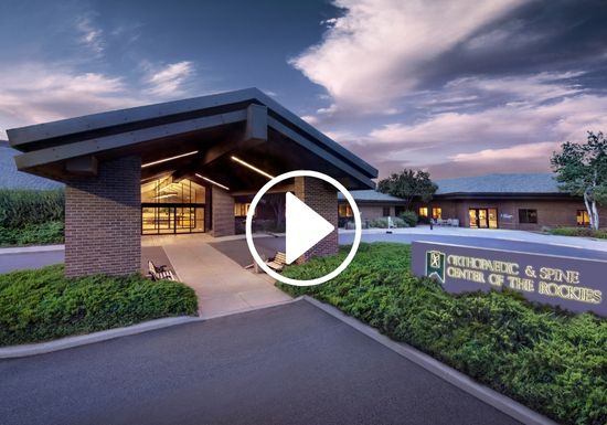Exterior view of the Orthopaedic & Spine Center of the Rockies building at dusk, with warm lights glowing inside, landscaped grounds, and a prominent entrance under a large overhang. A play button icon is overlaid on the image.