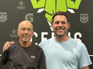Two men stand side by side, smiling, in front of a black wall with green and white gym logos and text. One, celebrating turning 70 and proudly embracing his shoulder comeback, wears a black shirt and blue shorts; the other wears a light blue shirt.